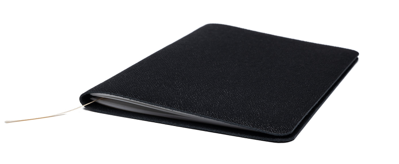 An angled view of the closed Pensift Notebook, highlighting the incredibly slim profile of the black Epsom leather cover and the subtle bookmark ribbon trailing from the pages.